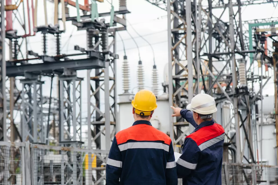 Two men in white and yellow hard hats and matching jackets looking up at power infrastructure in outdoor electrical substation or plant.